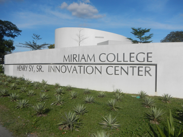 Außenansicht des Miriam College Innovation Center mit einer textbedeckten Wand, umgeben von Grün, einem Weg, Bäumen und einem bewölkten Himmel.