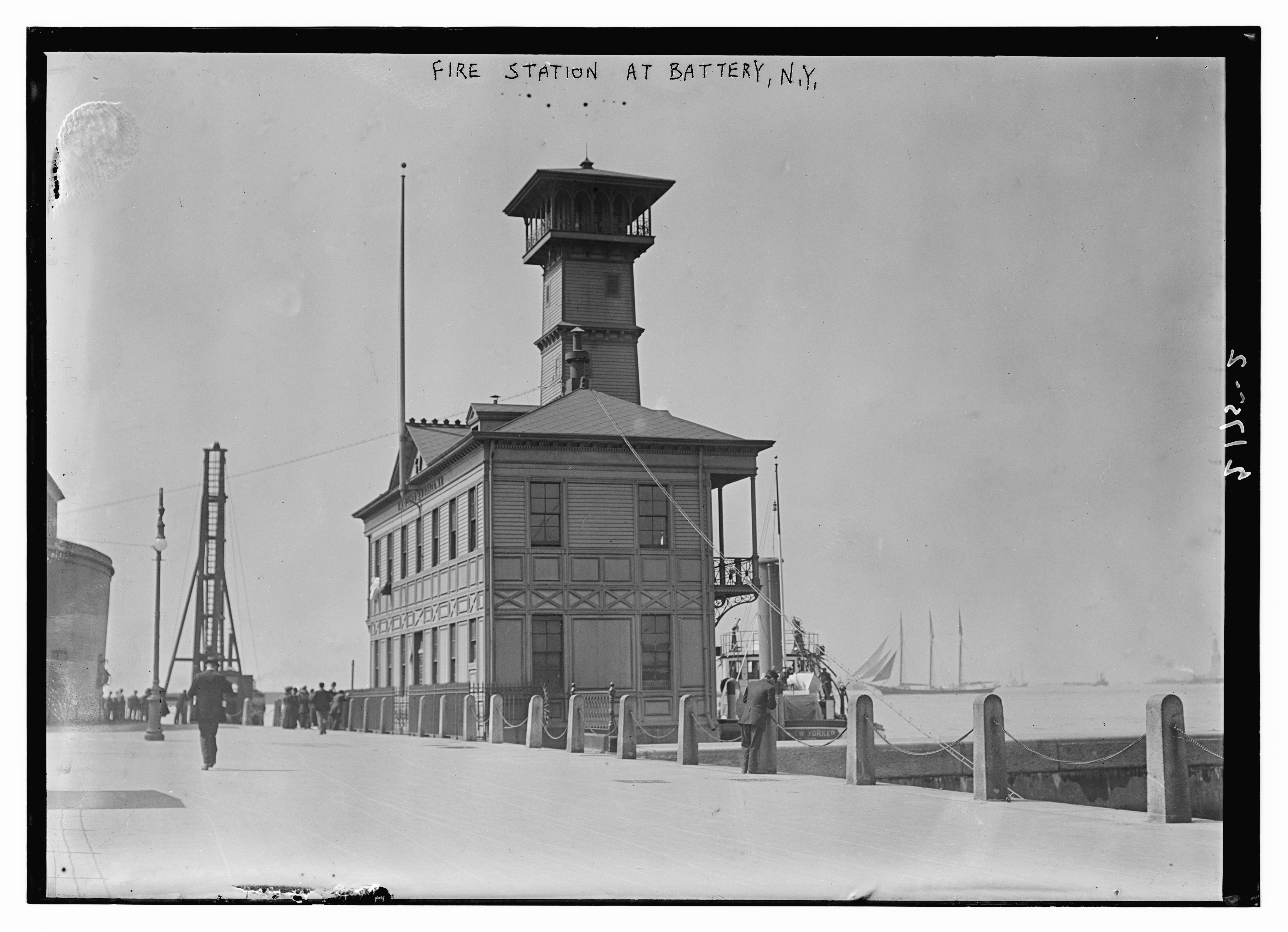 Ein Schwarz-Weiß-Foto einer Feuerwache in Battery, New York, mit einer Gruppe von Menschen auf dem Boden, Pfählen, einem Zaun, Booten auf dem Wasser und einem bewölkten Himmel.