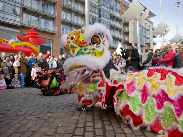 Ein lebendiges chinesisches Neujahrsfest in Amsterdam mit einer Löwendanzaustellung vor einer Zuschauermenge, einige halten Kameras, vor einem Hintergrund aus Gebäuden, Laternenmästen und einem klaren blauen Himmel.