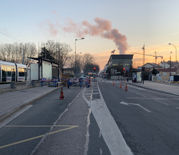 Eine Stadtstraße mit Verkehrskegeln, einem Bus, Laternen, Bäumen, Gebäuden, Kränen und Rauch, der aus einem entfernten Gebäude aufsteigt, unter einem Himmel.