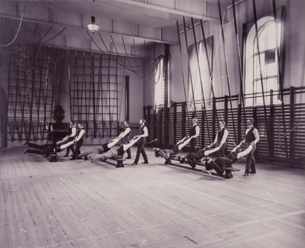 Schwarzes und weißes Bild einer Gruppe von Menschen, die Pilates-Übungen in einem Fitnessstudio mit einer Wand im Hintergrund, Fenstern auf der rechten Seite und Deckenleuchten durchführen.