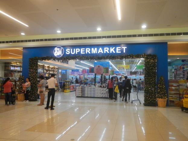 Gruppe von Menschen vor einem Supermarkt mit Einkaufstaschen, Einkaufswagen, Topfpflanze, Regalen, Schildern und einer Decke mit Leuchten.
