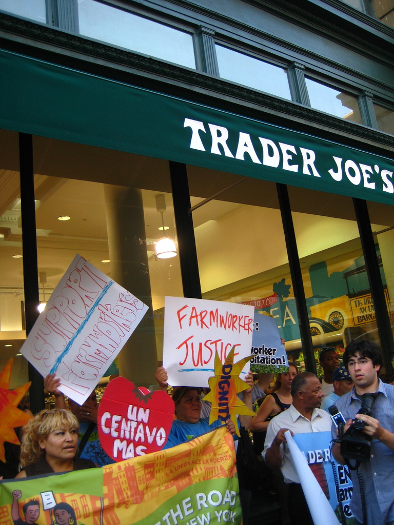 Eine Gruppe von Menschen steht in der Nähe eines Ladens und hält Schilder und Plakate hoch, wobei hinter ihnen eine Säule und Dachleuchten sichtbar sind.