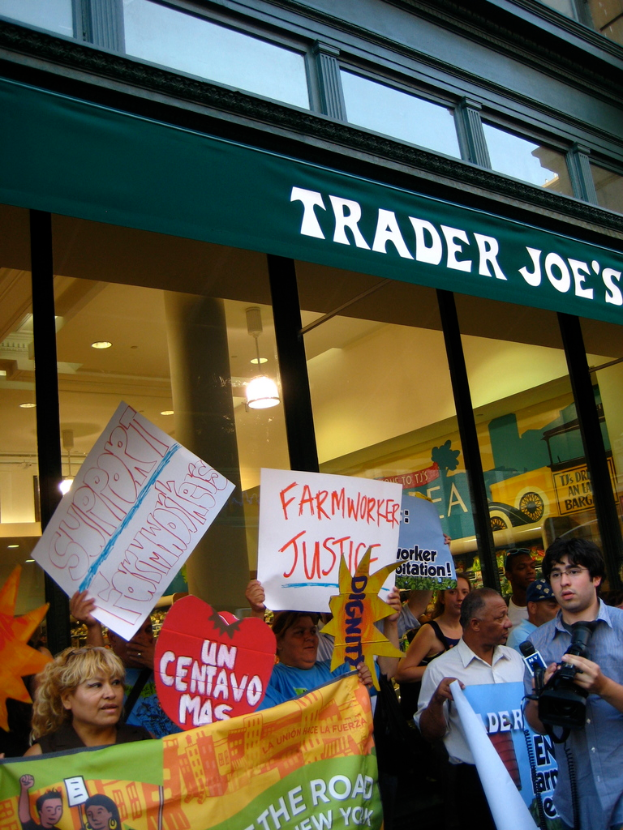 Eine Gruppe von Menschen steht in der Nähe eines Ladens und hält Schilder und Plakate hoch, wobei hinter ihnen eine Säule und Dachleuchten sichtbar sind.