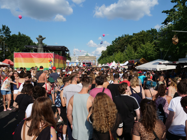 Eine große Menschenmenge geht eine Straße mit Zelten, Bäumen, Pfählen, Lichtern und einer Statue entlang, mit Gebäuden und einem bewölkten Himmel mit Ballons im Hintergrund während des Berliner Pride-Festivals.