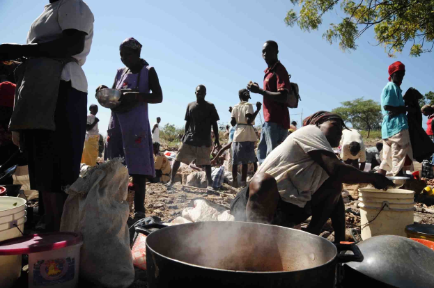 Eine Gruppe von Menschen, die in ländlicher Gegend Essen zubereiten, mit einem großen Topf, Eimern und Abdeckungen auf dem Boden, vor Bäumen und einem klaren blauen Himmel.