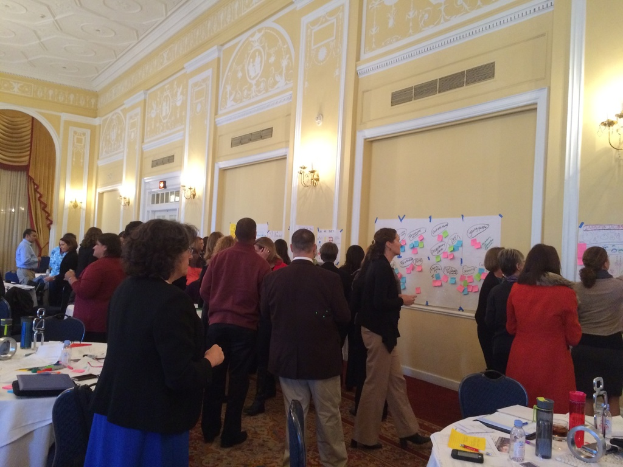 Eine Gruppe von Menschen untersucht Plakate und Diagramme, die an einer Wand in einem Raum zu sein scheinen, der wie ein Kunstmuseum aussieht.