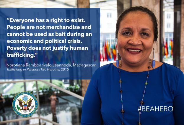 Eine Frau in einem blauen Kleid steht lächelnd vor einem Gebäude mit einer Aussage über Menschenhandel, mit Menschen, Pflanzen und Flaggen im Hintergrund.