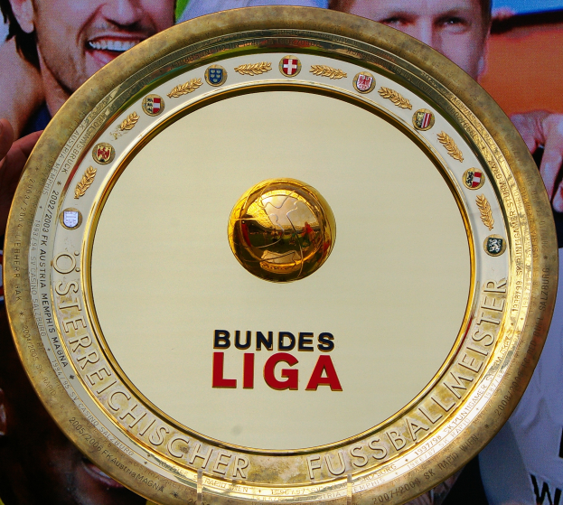 Mann hält goldenen Schild mit 'Bundesliga' hoch, im Hintergrund ein Banner mit drei Personen.