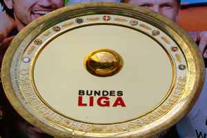 Mann hält goldenen Schild mit 'Bundesliga' hoch, im Hintergrund ein Banner mit drei Personen.