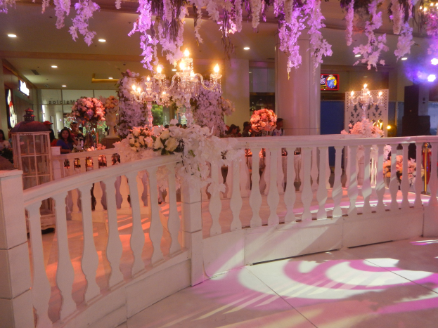 Weiße Reling mit floralen Dekorationen und hängenden Kronleuchtern bei einer Hochzeitsfeier, Menschen versammelt drumherum, beleuchtete Säulen im Hintergrund und ein Bildschirm auf der rechten Seite.