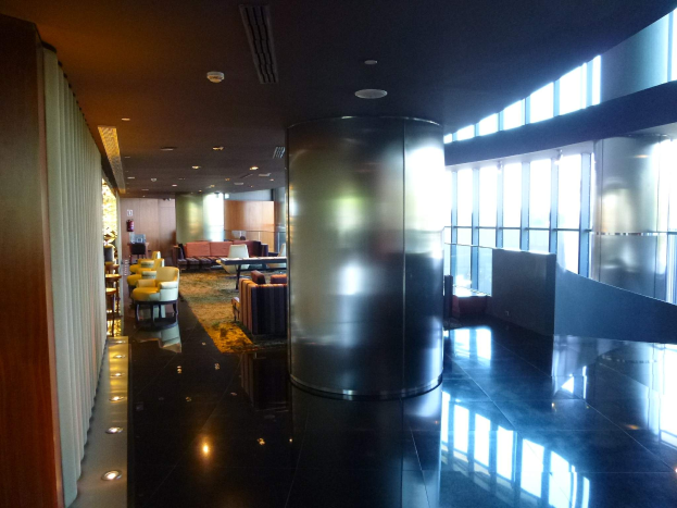Eine geräumige First-Class-Lounge am Hong Kong International Airport mit großen Fenstern, hohen Decken mit elektrischem Licht, Sitzanordnungen einschließlich Stühle und Sofas, Tischen, einem Teppichboden und einer zentralen Säule.
