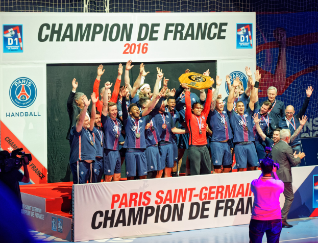 Eine Gruppe von Männern auf einer Bühne mit einem Pokal, einer Person, die filmt, und einem "Champion de France 2016"-Plakat im Hintergrund.