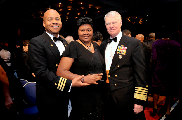 Zwei Männer und eine Frau stehen auf einem roten Teppich, die Frau hält einen Award, mit anderen im Hintergrund und einer beleuchteten Decke.