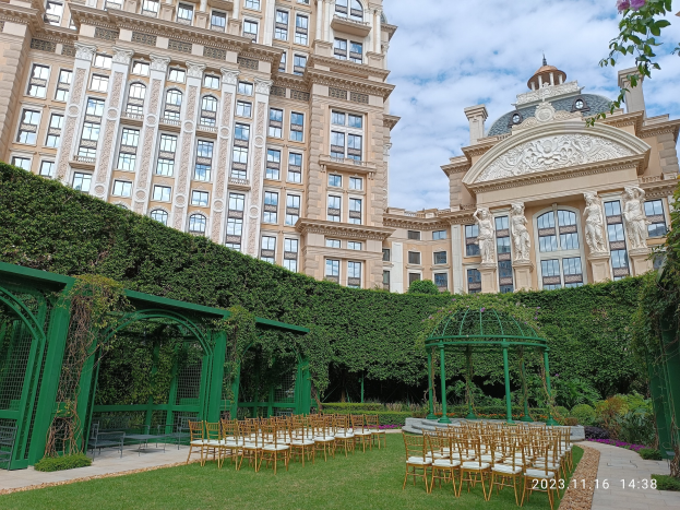 Hochzeit im Freien mit Stühlen, einem Pavillon, Grünpflanzen und einem großen Glasfensterbau unter einem blauen Himmel.