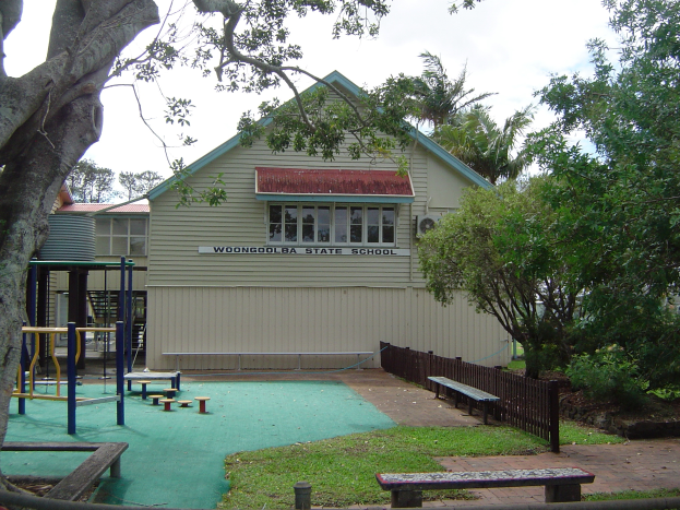 Ein Schulgebäude mit einem Spielplatz davor, umgeben von einem Zaun, Bänken, Gras, Pflanzen, Bäumen und anderen Gegenständen, unter einem bewölkten Himmel.