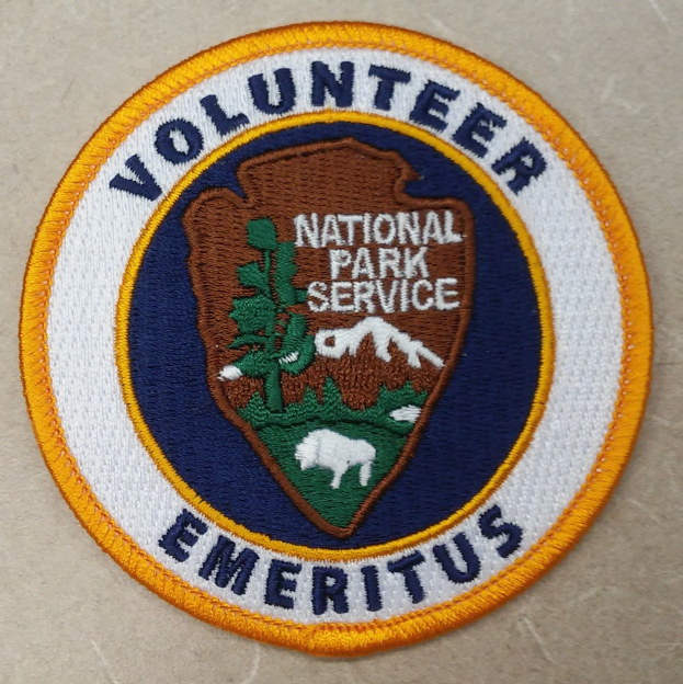 Runder Aufnäher mit blauem Rand und weißem Hintergrund, auf dem in schwarzer Schrift 'Volunteer National Park Service Emeritus' in einem gelben Kreis steht.