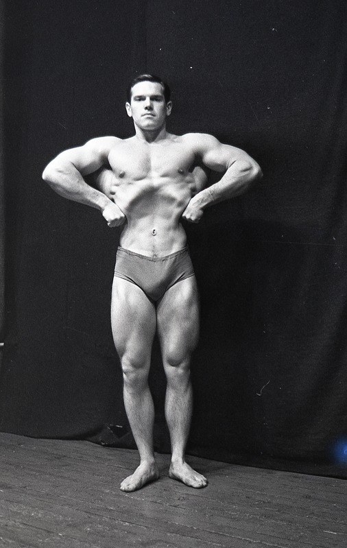 Bodybuilder auf einer Bühne mit Händen in den Hüften stehend, in Schwarz-Weiß-Kleidung, mit einem Vorhang im Hintergrund.