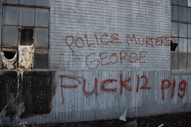 Ein Gebäude mit Graffiti, das "Polizei Mord George Fuck 12 P19" auf der Seite trägt, mit Fenstern und verschiedenen Gegenständen davor.