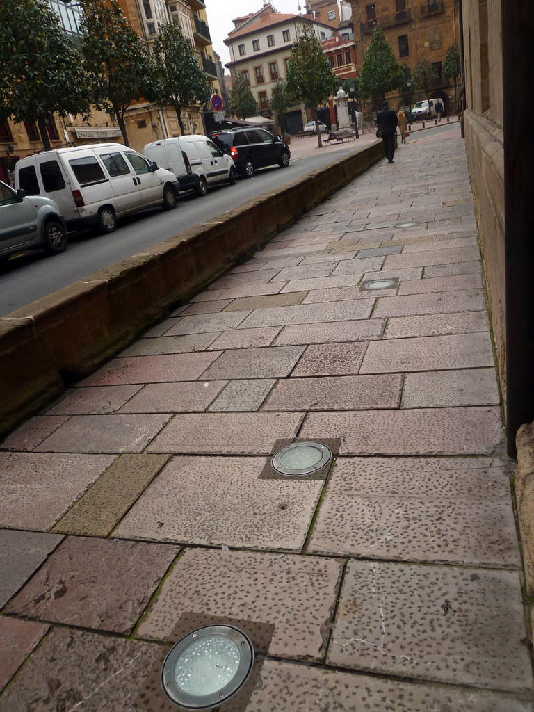 Mehrere Fahrzeuge stehen auf einer Straße mit einer Person auf dem benachbarten Gehweg, Häuser, Bäume und Strommästele im Hintergrund bei klarem Himmel.