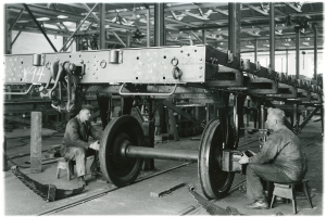 Zwei Männer sitzen auf Hockern in einer Fabrik und arbeiten an einem Waggon mit Werkzeugen, umgeben von Metallobjekten, Stäben und Deckenlampen, auf einem Schwarz-Weiß-Bild.