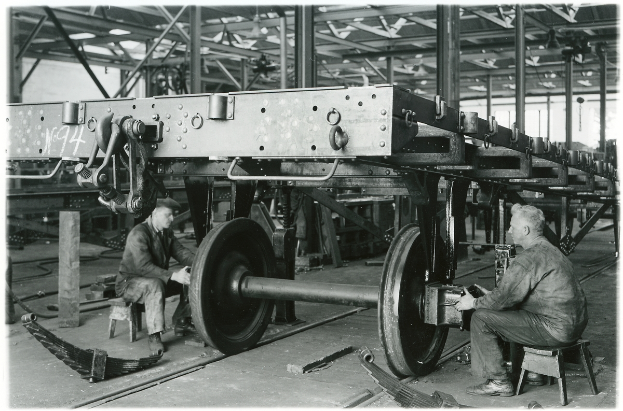 Zwei Männer sitzen auf Hockern in einer Fabrik und arbeiten an einem Waggon mit Werkzeugen, umgeben von Metallobjekten, Stäben und Deckenlampen, auf einem Schwarz-Weiß-Bild.