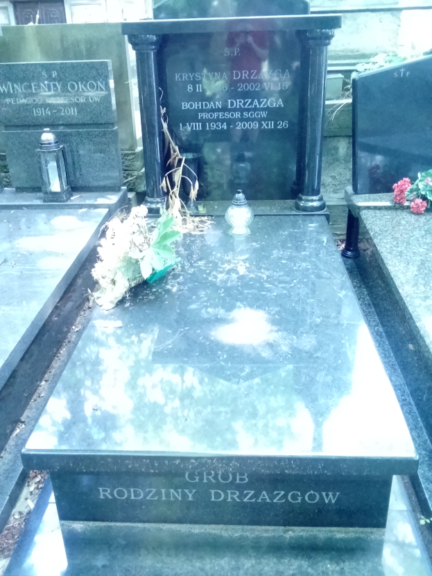 Ein Friedhof mit zwei benachbarten Grabsteinen umgeben von Blumenbouquets und anderen Gegenständen, mit einem Gebäude im Hintergrund.