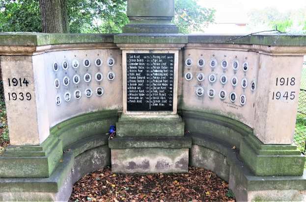 Ein Holocaust-Gedenkmonument in einem jüdischen Friedhof in Berlin, das eine Tafel mit Text und Zahlen zeigt, umgeben von Bäumen, einem Zaun und verstreuten trockenen Blättern.