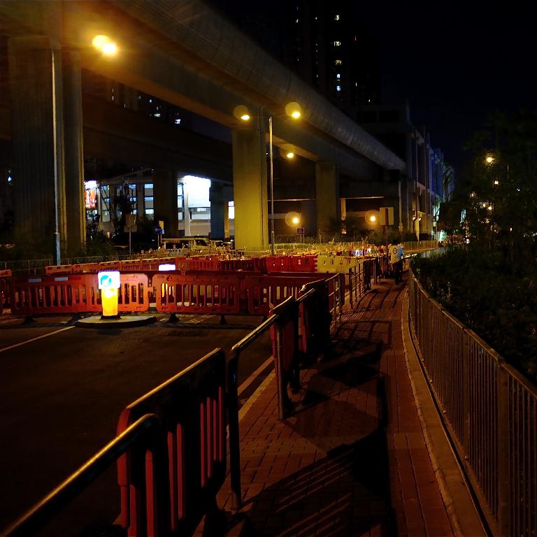 Eine nächtliche Szene mit roten Zaunlatten auf einer Straße, Bäumen, Gebäuden, Lichtern im Hintergrund und einer Brücke in der Ferne sowie einem weiteren Zaun auf der rechten Seite.