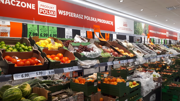 Ladeninneres mit verschiedenen Obst- und Gemüsesorten in Kartonschachteln, jede mit Preisschild, Wandtafeln mit Text und Bildern und Deckenbeleuchtung.