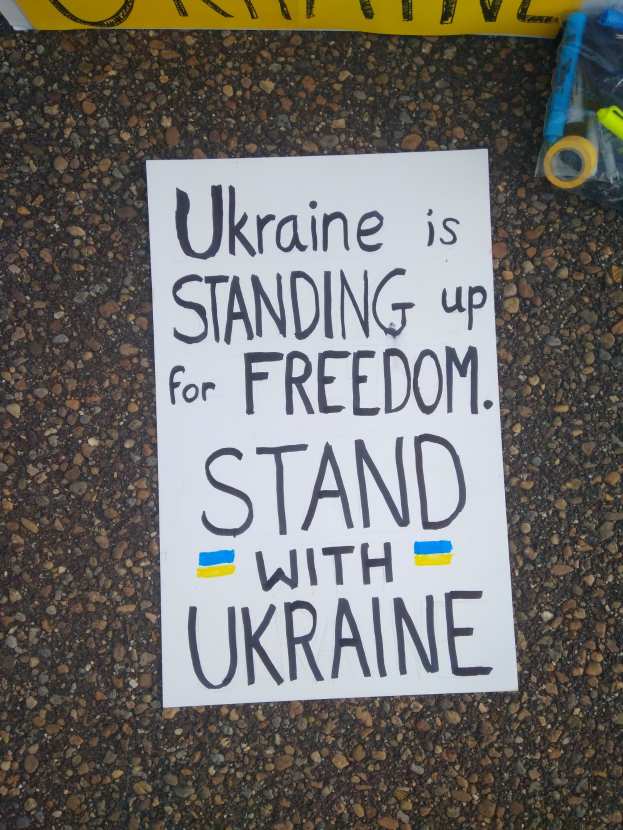Weißer Papierbogen mit "Ukraine steht für Freiheit, steht mit Ukraine" darauf vor einem gelben Banner, mit einer Plastikhülle mit verschiedenen Gegenständen auf der rechten Seite.