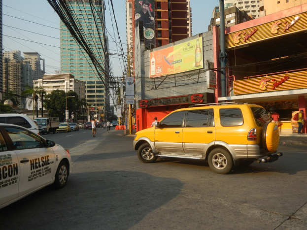 Ein gelbes Taxi fährt auf einer Stadtstraße mit hohen Gebäuden, Fußgängern auf dem Gehweg, Strommasten mit Drähten, Bäumen am Straßenrand und einem sichtbaren Himmel im Hintergrund.