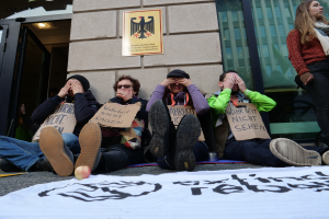 Eine Gruppe von Menschen sitzt vor einem Gebäude auf dem Boden und hält Schilder hoch, mit einer Fahne und einem Apfel auf der Straße, und eine Frau steht rechts daneben und sieht besorgt aus.