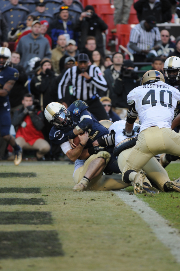 Ein Fußballspieler wird von einer Gruppe von Verteidigern auf einem Rasenfeld getackelt, mit Zuschauern und Kameramännern im Hintergrund, alle Spieler tragen Helme.