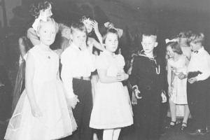 Eine Gruppe von Kindern, die zusammen auf einer Tanzfläche stehen und fröhlich feiern, mit einer Wand im Hintergrund, dargestellt in Schwarz-Weiß.