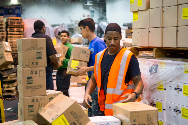 Eine Gruppe von Männern in orangefarbenen Westen und Sicherheitswesten lädt Kisten in ein Lagerhaus, mit mehreren Kartonagen auf der rechten Seite und einer Kette auf der linken Seite und einer beleuchteten Wand im Hintergrund.