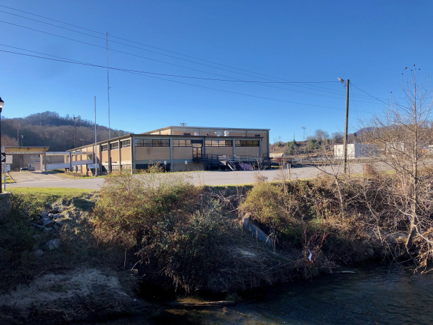 Grüne Wiese mit einem Fluss, Bäumen, Pflanzen, Gras, Steinen, Straßenlaternen, Strommasten, Stromkabeln, Hügeln, einem klaren blauen Himmel und einem Gebäude, das für eine Wasseraufbereitungsanlage geplant ist.