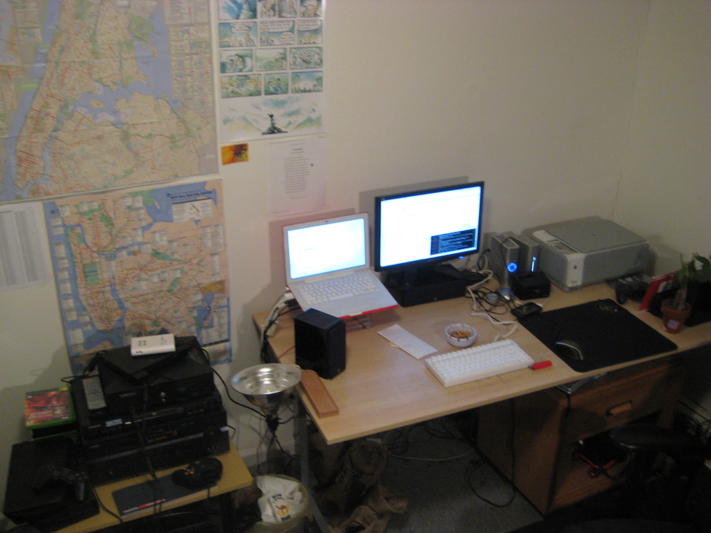 Ein Raum mit einem Tisch, auf dem ein Laptop, ein Lautsprecher, ein Monitor, eine Tastatur, ein Ball, eine Maus, ein Scanner und Papiere stehen; ein weiterer Tisch mit Elektronikgeräten in der linken unteren Ecke; und eine Wand mit Charts und Postern im Hintergrund.