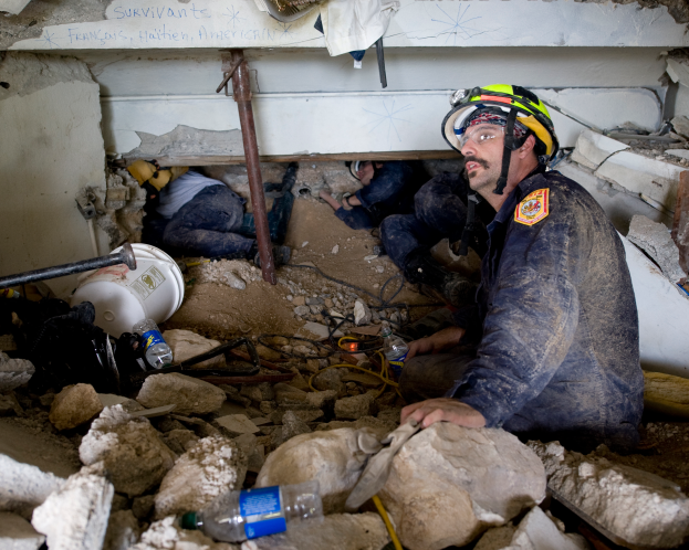 Ein Feuerwehrmann mit Helm sitzt zwischen Trümmern, darunter Steine und Flaschen, mit einer Wand im Hintergrund, auf der ein Text zu sehen ist.