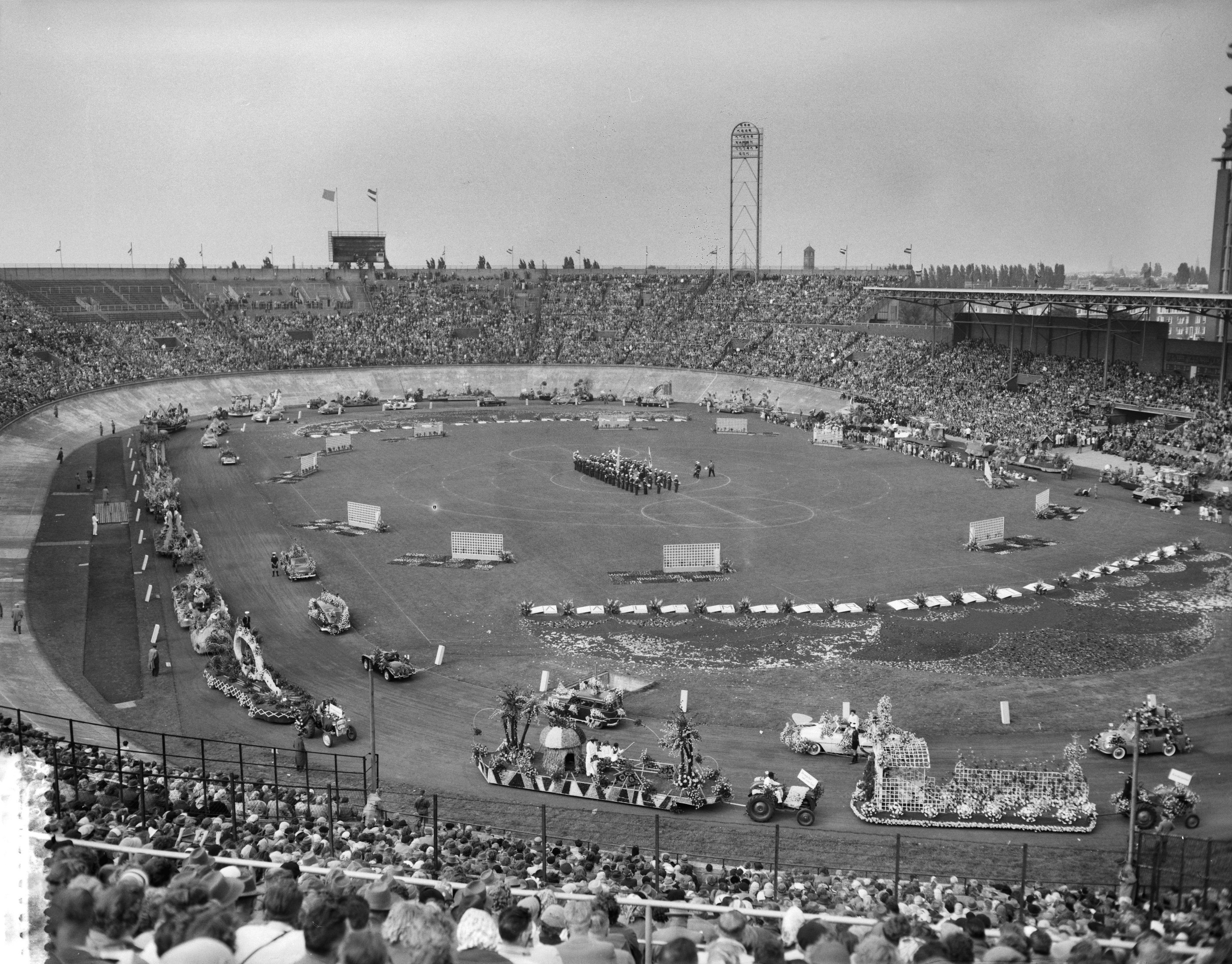 Schwarzes und weißes Bild einer großen Menschenmenge, die ein Rennen in einem Stadion mit Fahrzeugen, Zuschauern, Absperrungen, Pfosten, Fahnen, Türmen, Bäumen und Himmel sieht.