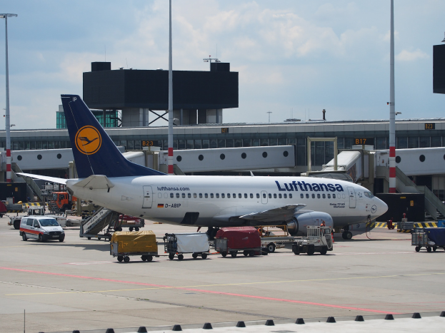 Lufthansa Airbus A320-200-Flugzeug auf dem Frankfurter Flughafen geparkt mit Bodenfahrzeugen und Wagen im Vordergrund, einem Terminalgebäude, Laternenmasten und einem klaren blauen Himmel im Hintergrund.