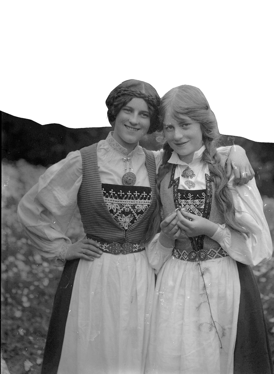 Zwei junge Frauen in traditioneller bayerischer Tracht, mit weißen und schwarzen Kleidern mit aufwendiger Stickerei und Perlen, posieren vor einer Kulisse aus üppigen grünen Pflanzen und einem hellen Himmel.