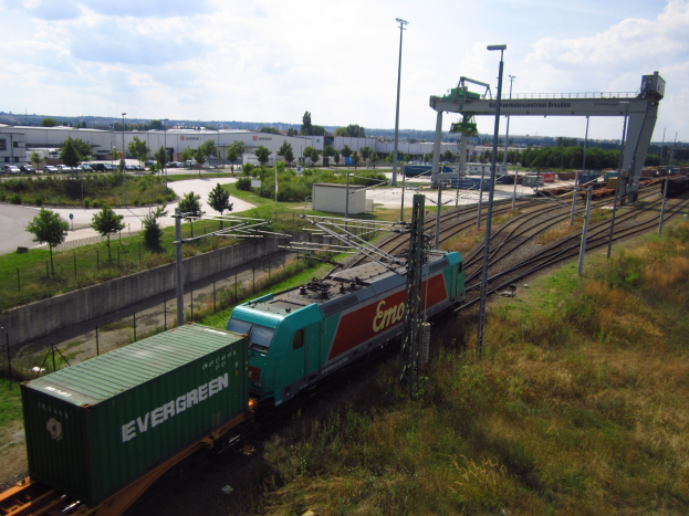 Güterzug fährt auf Schienen neben einem grünen Feld mit Strommasten, Kabeln, Gras, Bäumen, Gebäuden, Fahrzeugen auf der Straße und einem bewölkten Himmel.