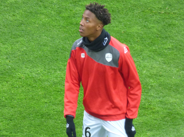 Ein junger Mann in roter Jacke und weißen Shorts steht auf einem Fußballfeld, trägt Baseballhandschuhe und schaut entschlossen nach rechts.
