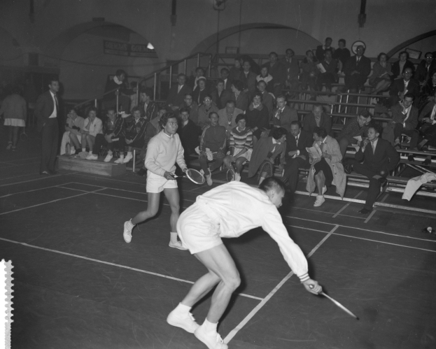Zwei Männer spielen Badminton auf einem Court, mit einer Zuschauermenge auf Tribünen sitzend und anderen stehend, mit einer Wand im Hintergrund.