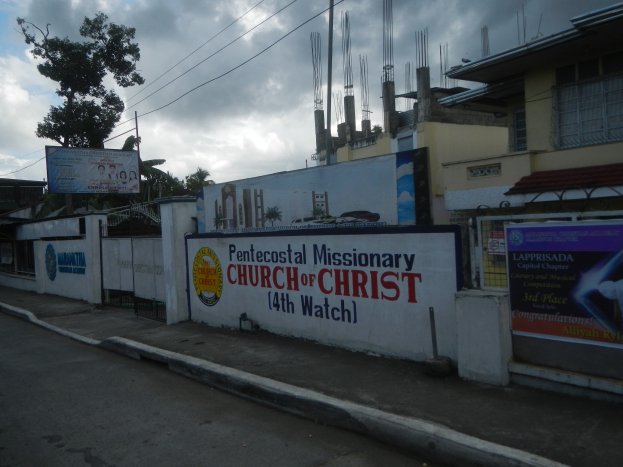 Die Pfingstmissionarische Kirche Christi 14. Wacht steht mit einer Straße im Vordergrund, einer Wand mit Bannern und Text, Gebäuden, Bäumen, Masten, Drähten und einem bewölkten Himmel im Hintergrund.