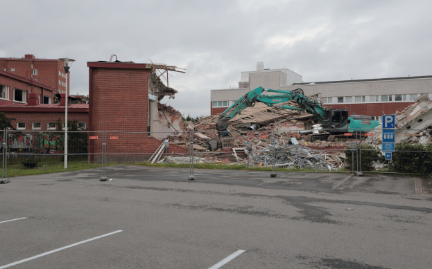 Großes Gebäude in einem Parkplatz mit einem Bagger, Metallstangen, Zaun, Schild, Pflanzen, Gras, Bäumen, benachbarten Gebäuden und bewölktem Himmel.