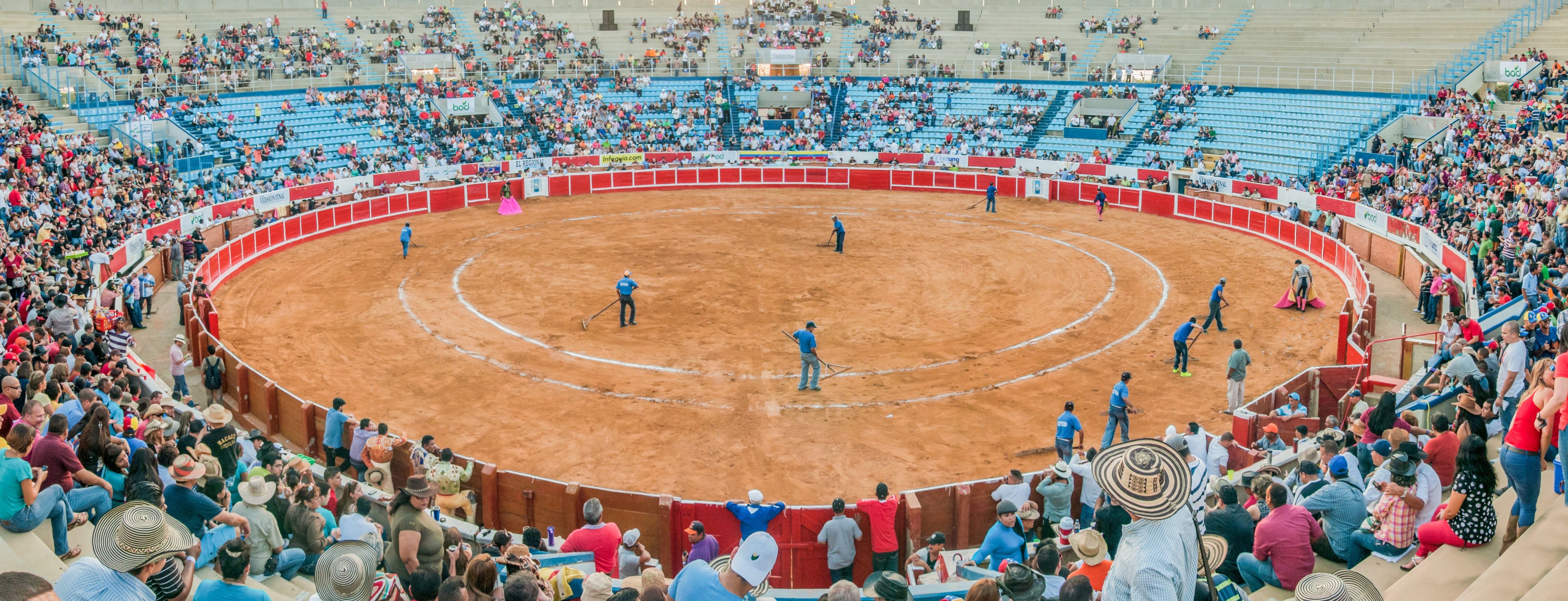 Eine große Menschenmenge schaut einer Stierkampfveranstaltung in einem Stadion zu, wobei einige Menschen stehen, sitzen oder Gegenstände halten und eine Person unten einen Hut trägt.