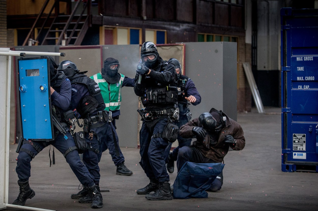 Eine Gruppe von Polizisten in Einsatzausrüstung steht vor einem Gebäude in einer Reihe, wobei eine Person im Vordergrund liegt und ein blaues Objekt mit Text auf der rechten Seite zu sehen ist.