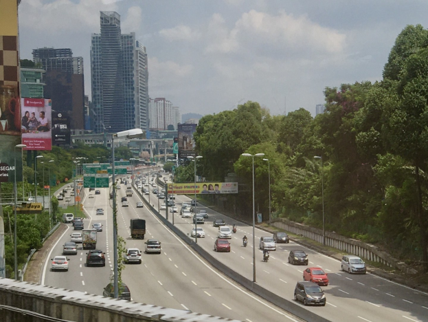 Eine belebte Autobahn mit hohen Gebäuden, Straßenlaternen, Werbetafeln, Bäumen und einem bewölkten Himmel.